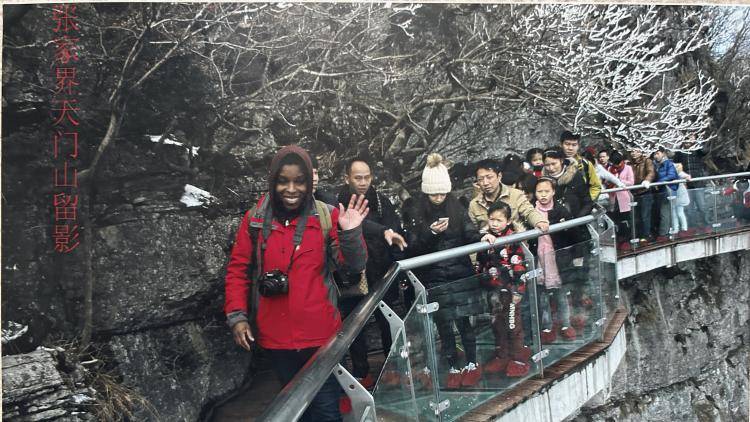 The writer Noo Saro Saro-Wiwa waves at the camera from a suspended walkway on a cliffside in China, with other tourists behind her