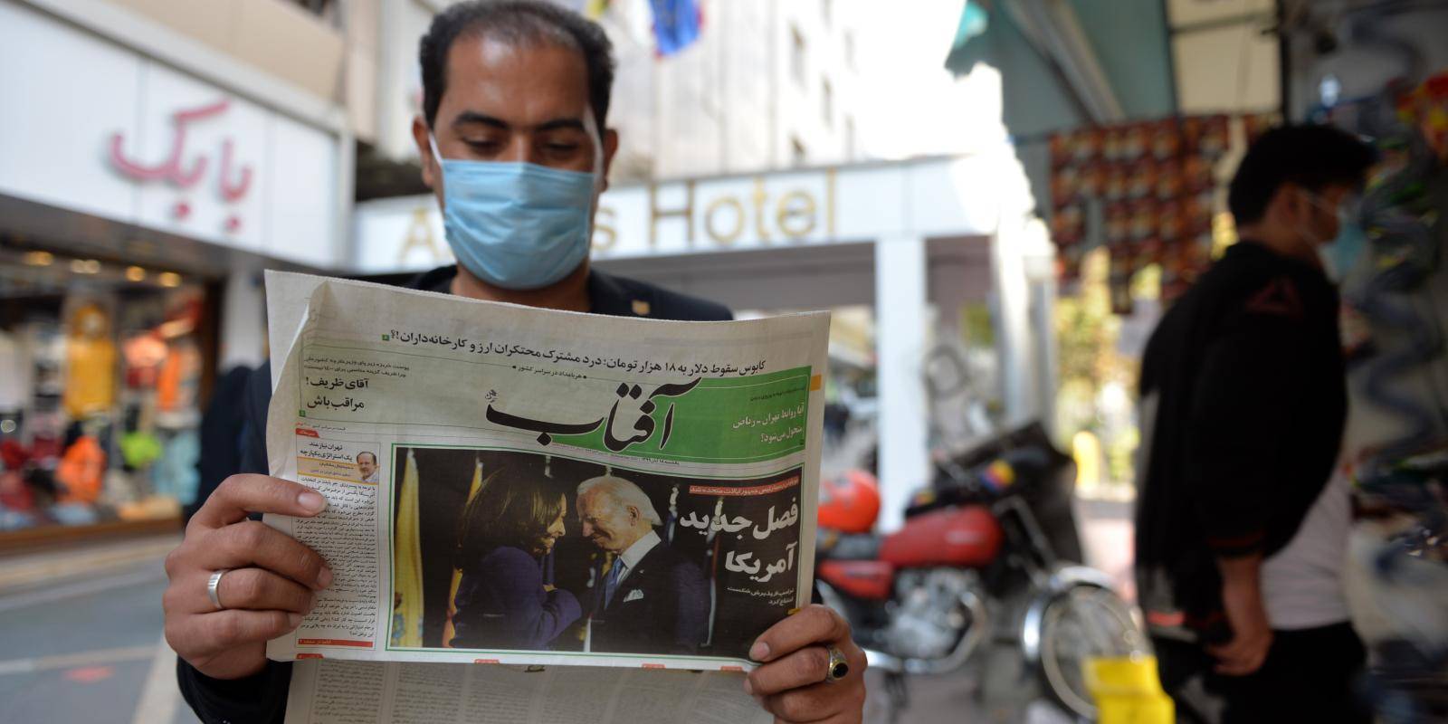A man stands on a Tehran street reading a Farsi-language newspaper showing a picture of Joe Biden