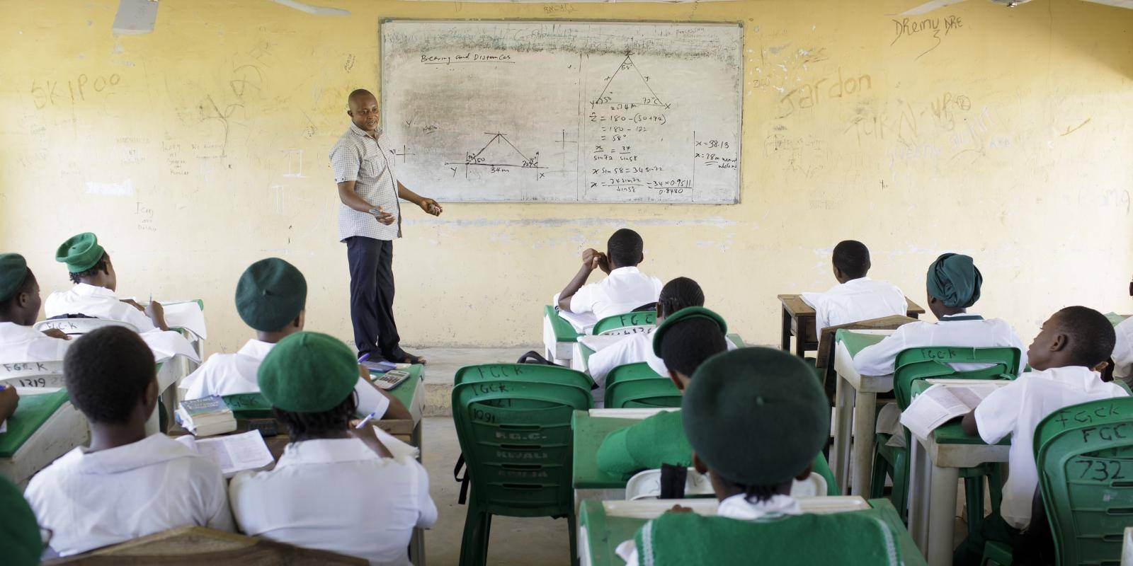Photo shows a teacher in a Nigerian high school standing by a whiteboard with a pen in his hand. Pupils are facing away from the camera watching him