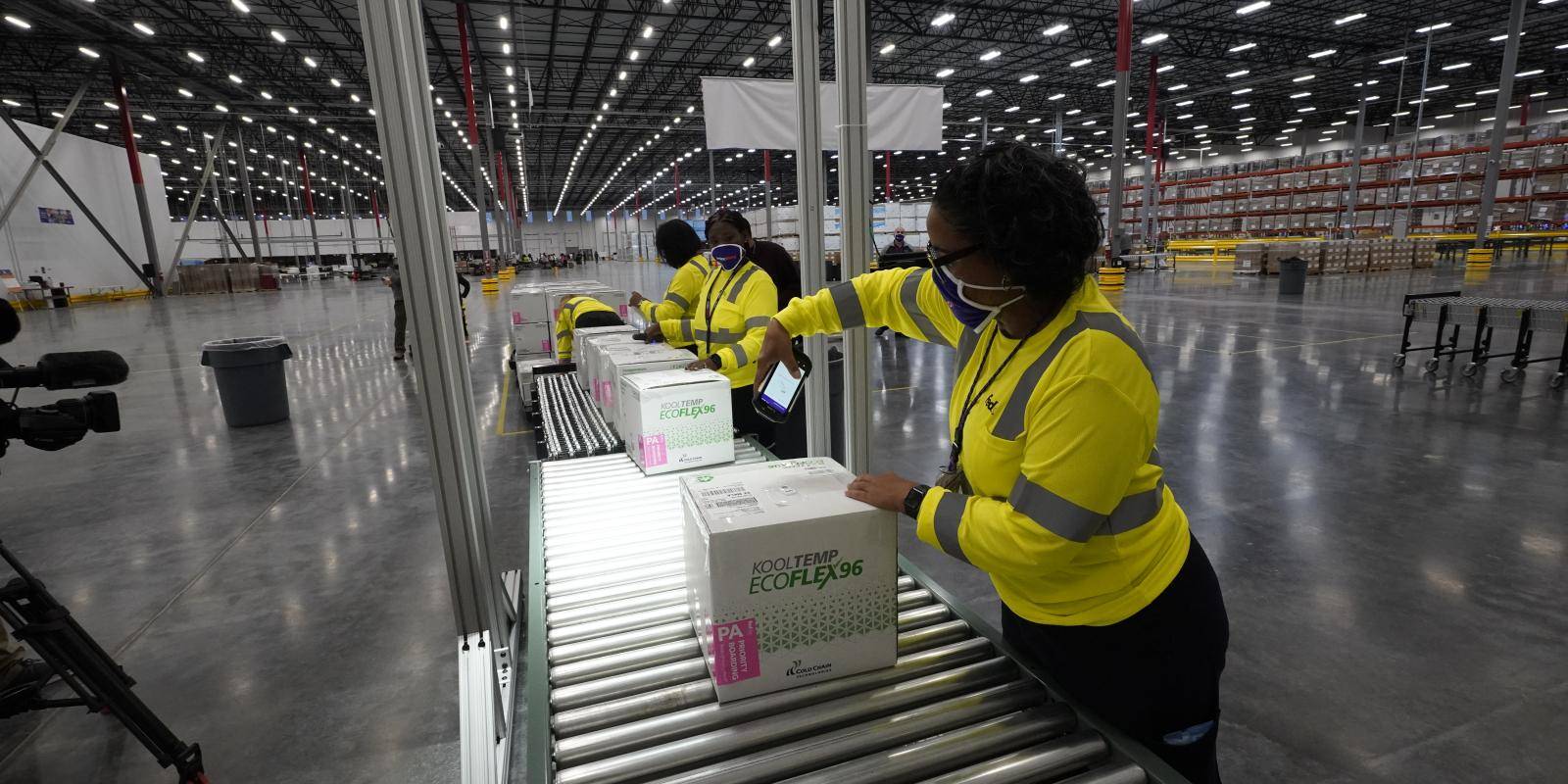 Photo shows a view along a production line where several masked workers are packing boxes of the Moderna COVID-19 vaccine