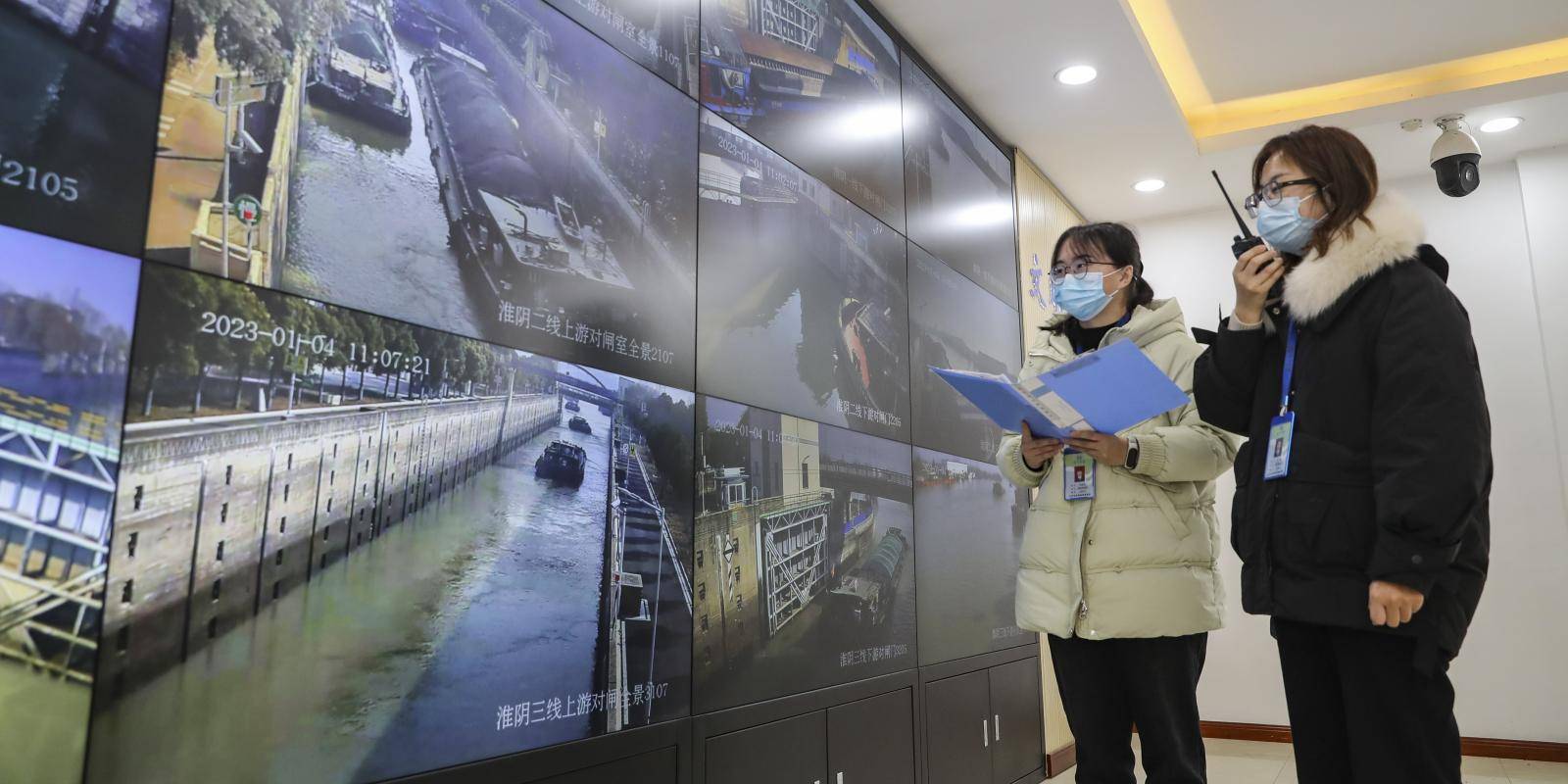 Two women dispatch administrators in warm coats and face masks stand in front of monitors showing images of transport barges on water.
