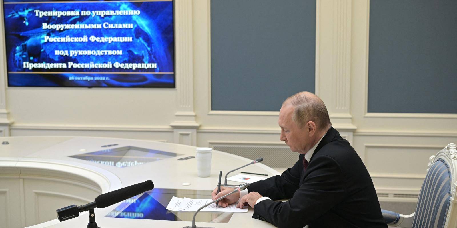 Russian president Vladimir Putin sits at a light-coloured desk writing while a wall-mounted video screen shows a link to a military training exercise