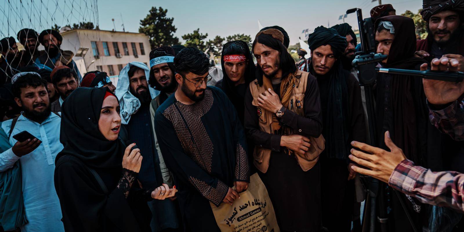 A female journalist talks to camera while standing in front of a crowd of male supporters and members of the Taliban.