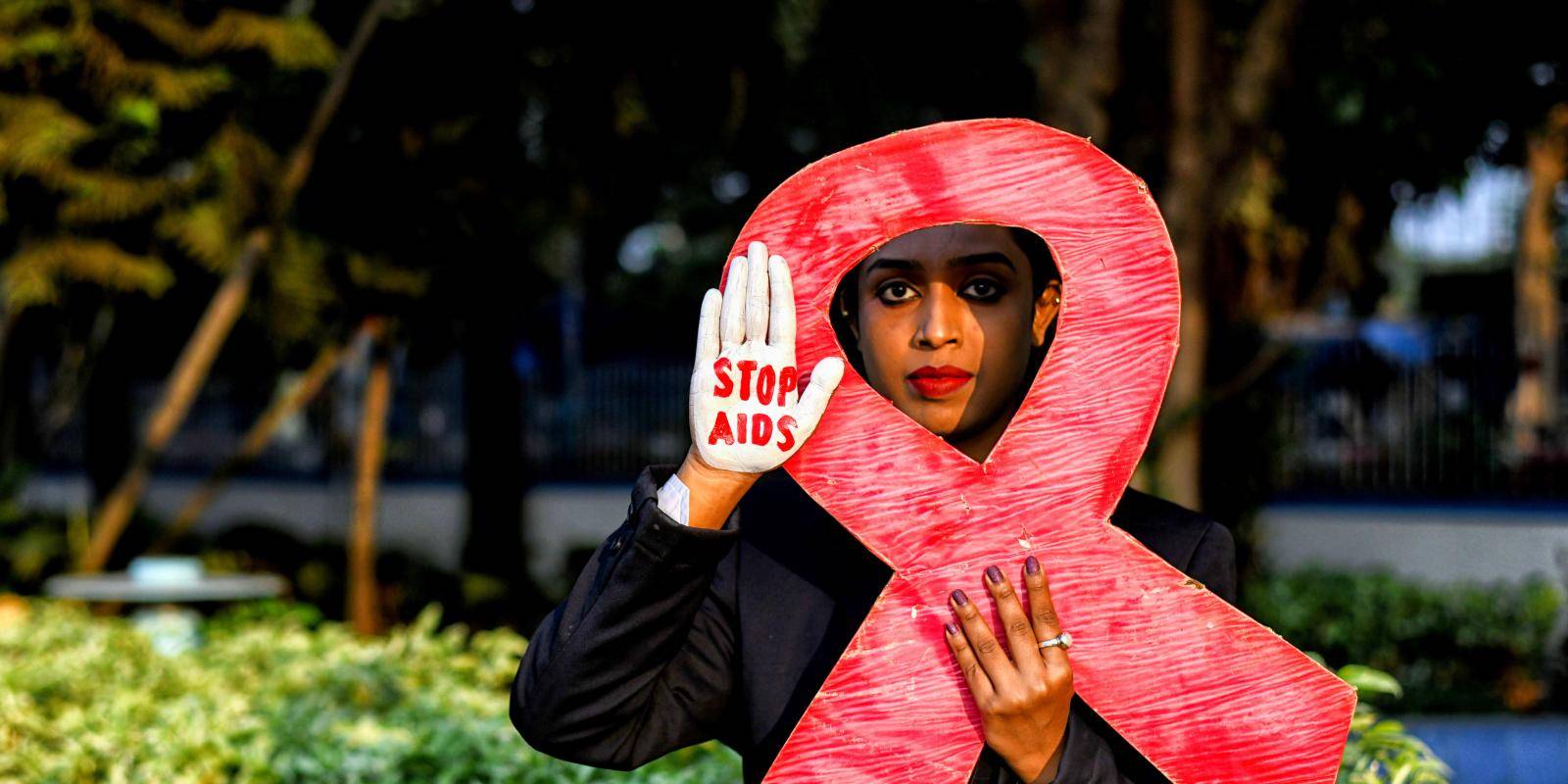 An activist dressed in black holds a replica of the red ribbon symbolizing awareness of and support for those living with HIV. They also raise the palm of their right hand, which is painted with the slogan 'Stop AIDS'.