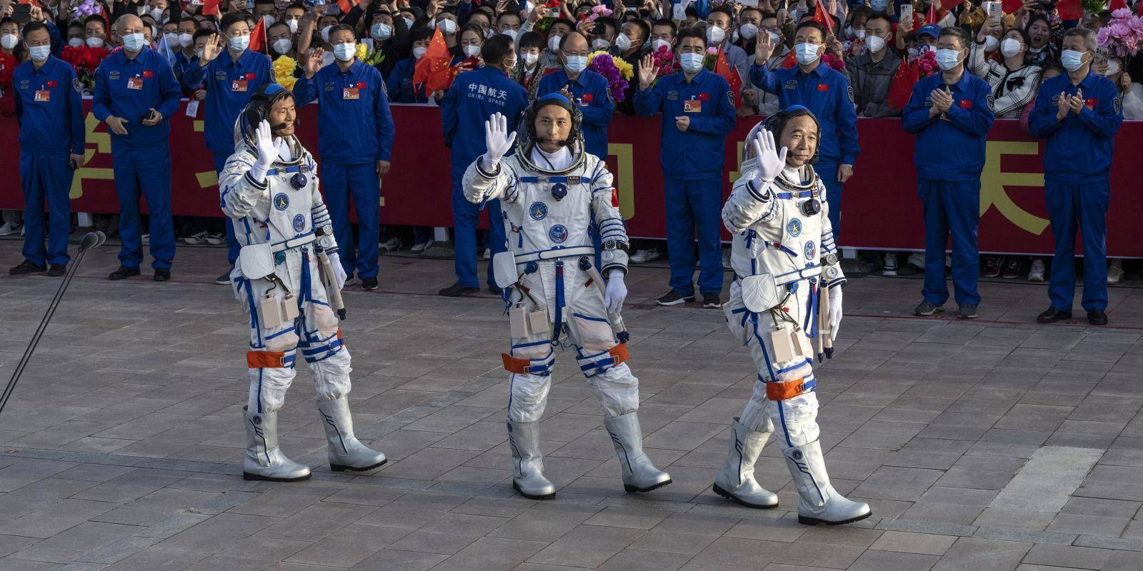 Three men in astronaut suits waving in front of a large crowd