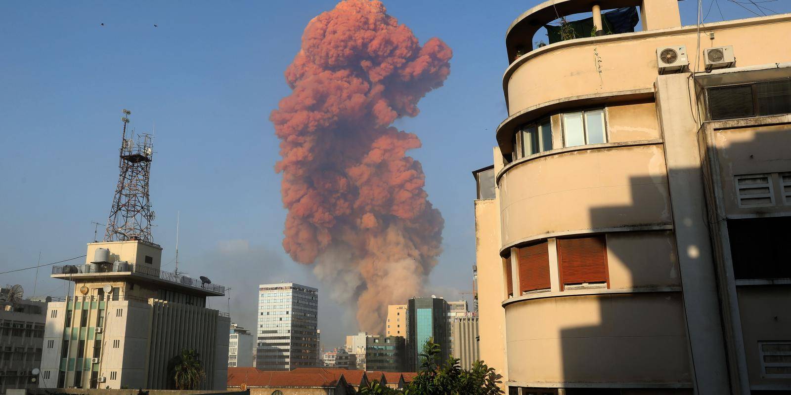 A large red-coloured plume of smoke rises into a blue sky above tall buildings and a telecommunications mast after an explosion.