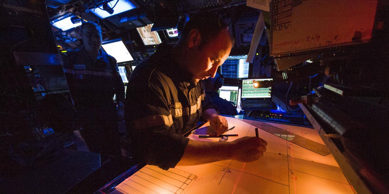 A man in naval uniform sits at a desk in front of illuminated screens onboard a submarine.