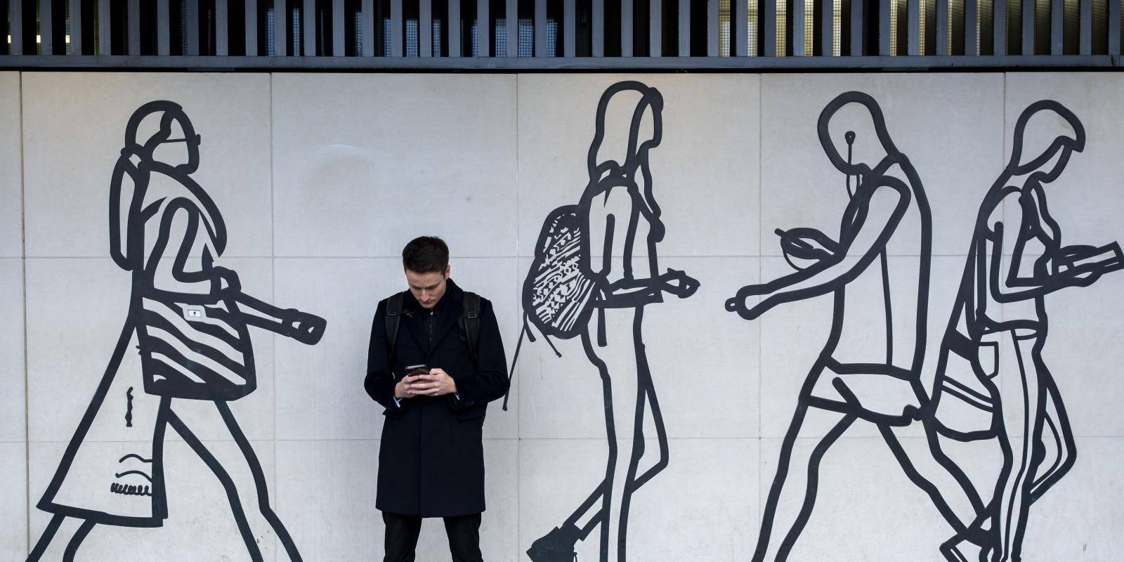 A man stands holding his mobile phone, in front of a mural drawn in black and white of people walking and using mobile devices.