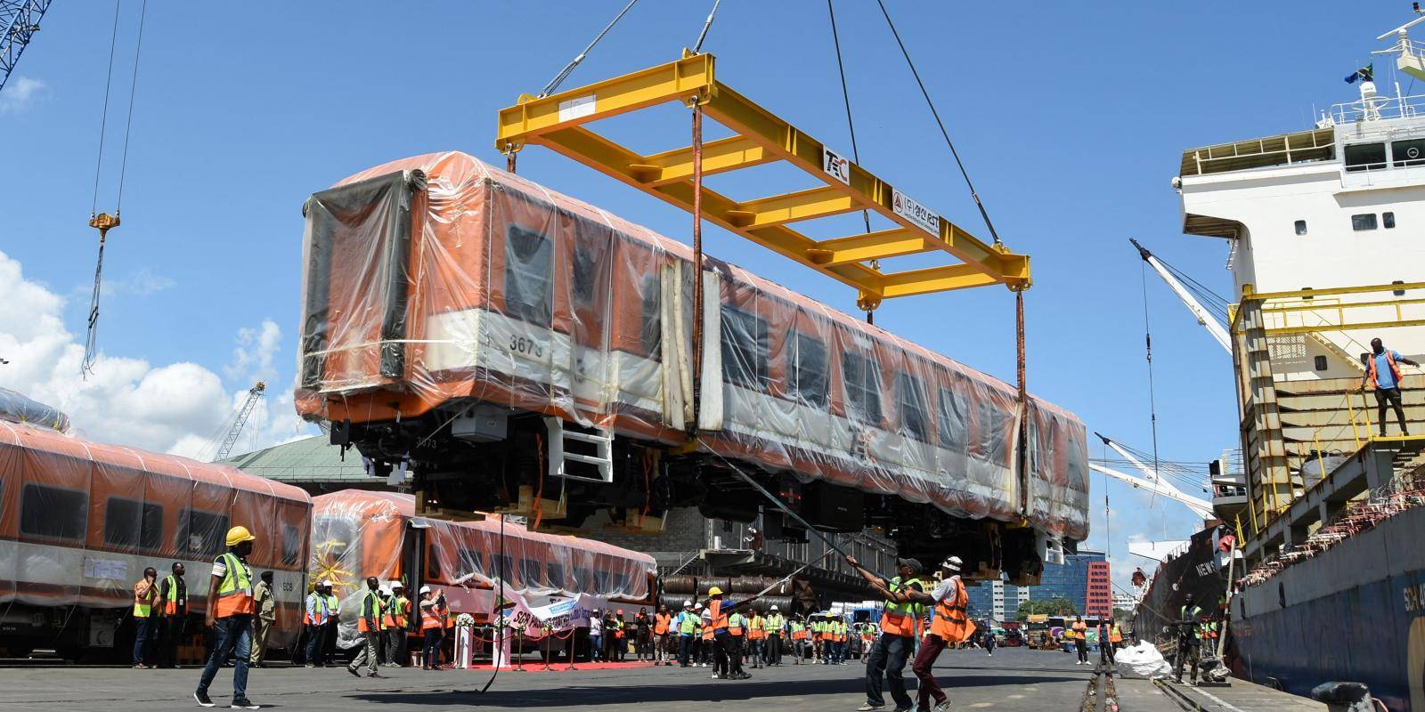 A brand-new train carriage hangs from a crane as workers in high-visibility clothing stand below use ropes to unload it from a ship onto the quayside.