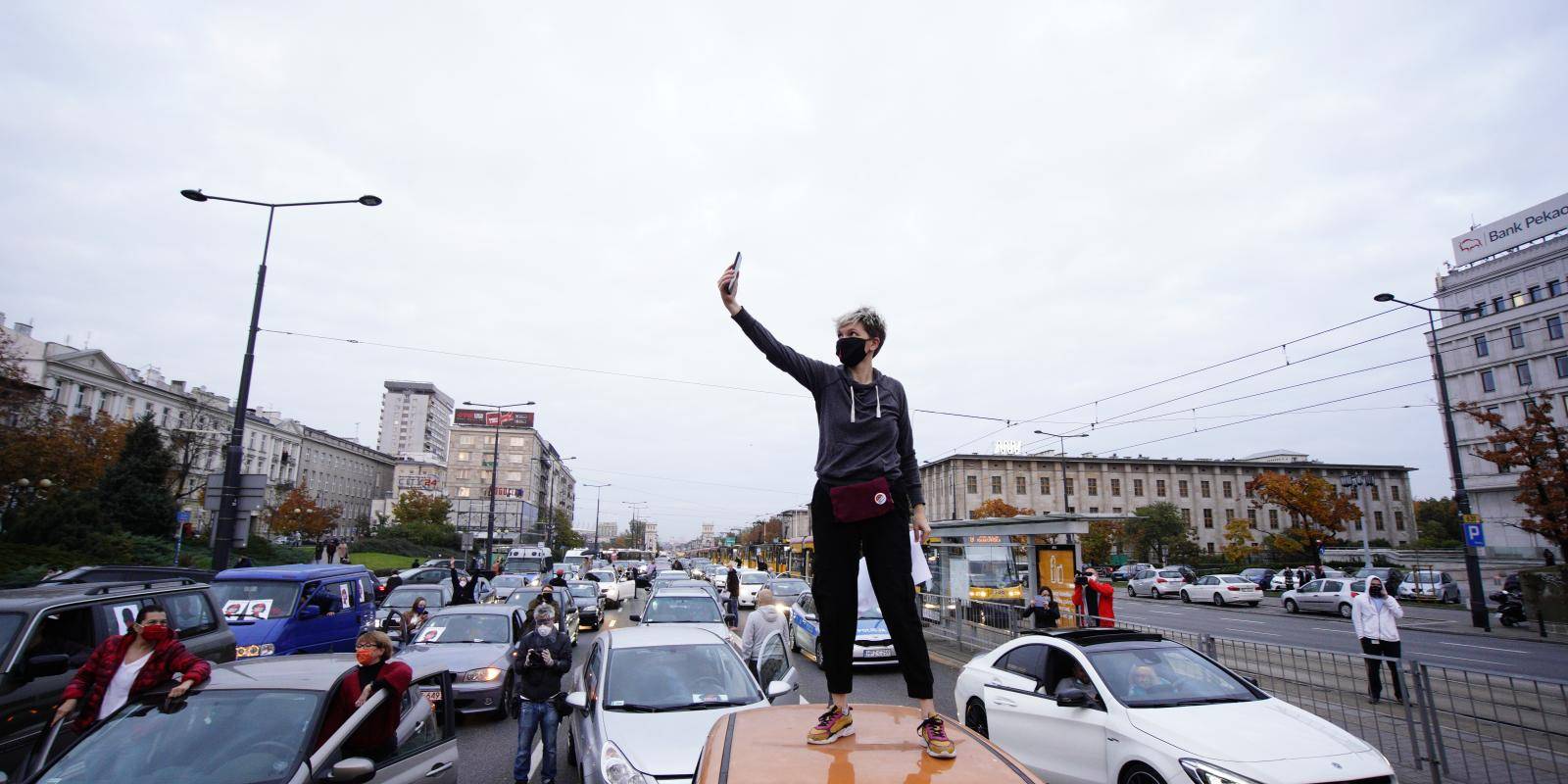 A woman in dark leisurewear and face mask taking a selfie on top of an orange car