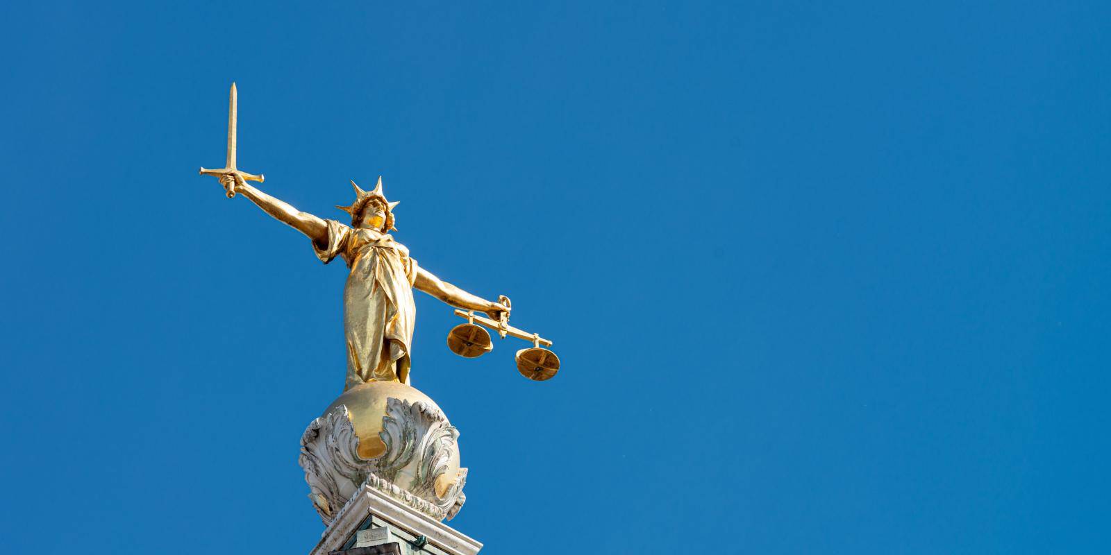 A statue of Lady Justice holding a sword and balancing scales on a sky blue background on top of the Old Bailey.
