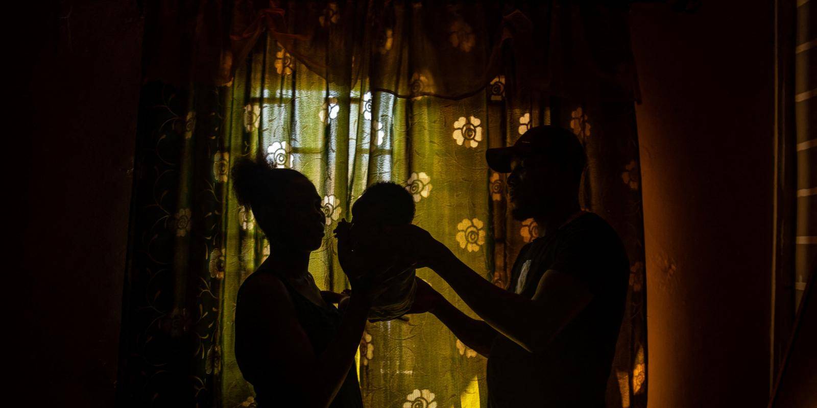 A couple stand facing each other in front of a green curtain, holding a baby between them.