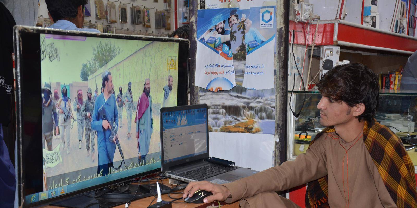 A young person sits at a table in a shop watching a screen showing images of Taliban fighters.