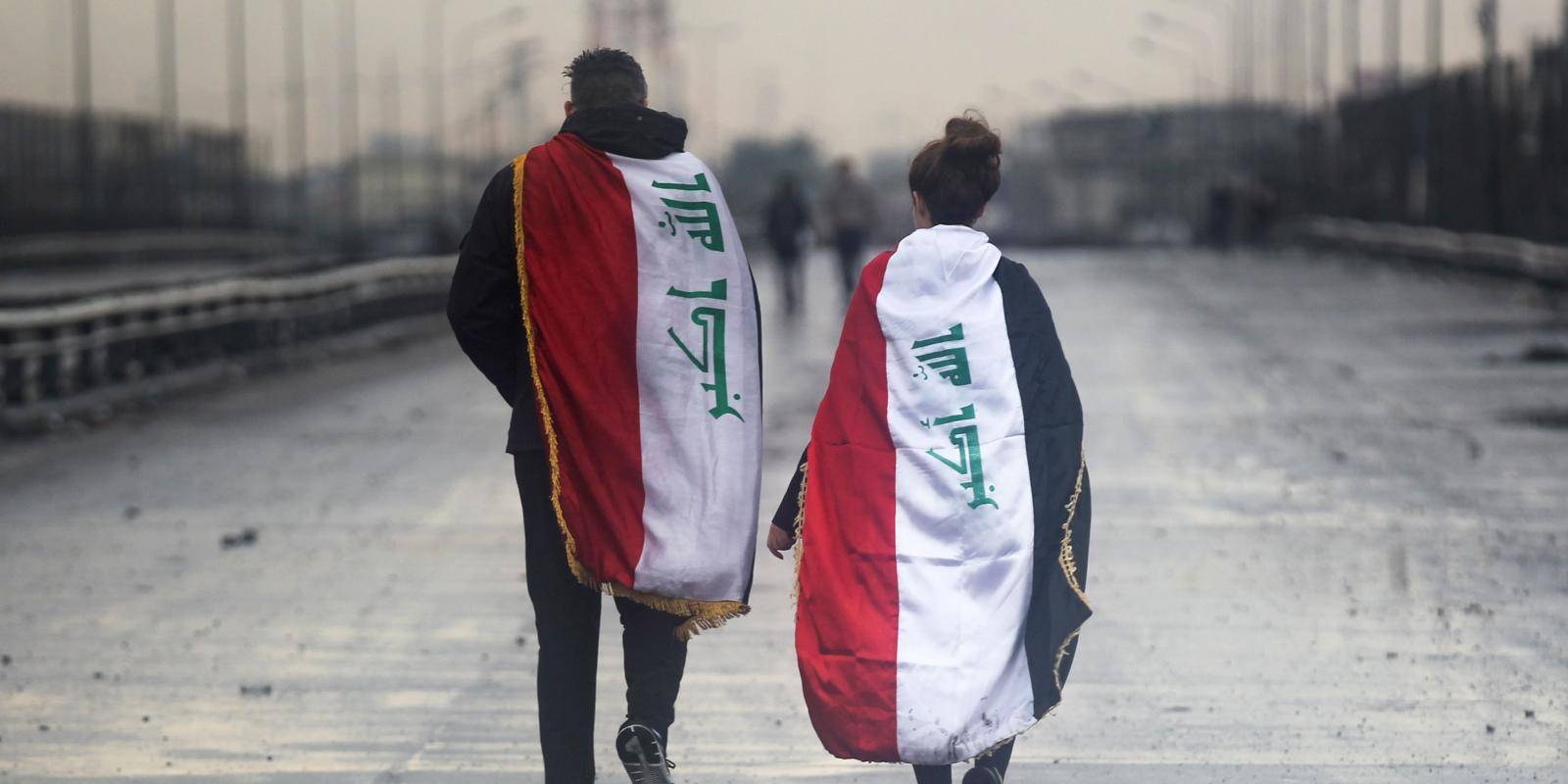 A young man and woman wearing Iraqi national flags on their backs walk away from the camera on a highway in Baghdad