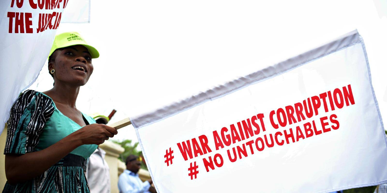 A woman dressed in blue and green holds a white flag with the protest slogan #war against corruption #no untouchables in ret capital letters