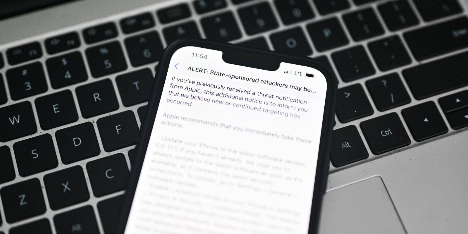 An iPhone held above a Mac keyboard displays an Apple alert saying state sponsored attackers may be targeting your iPhone