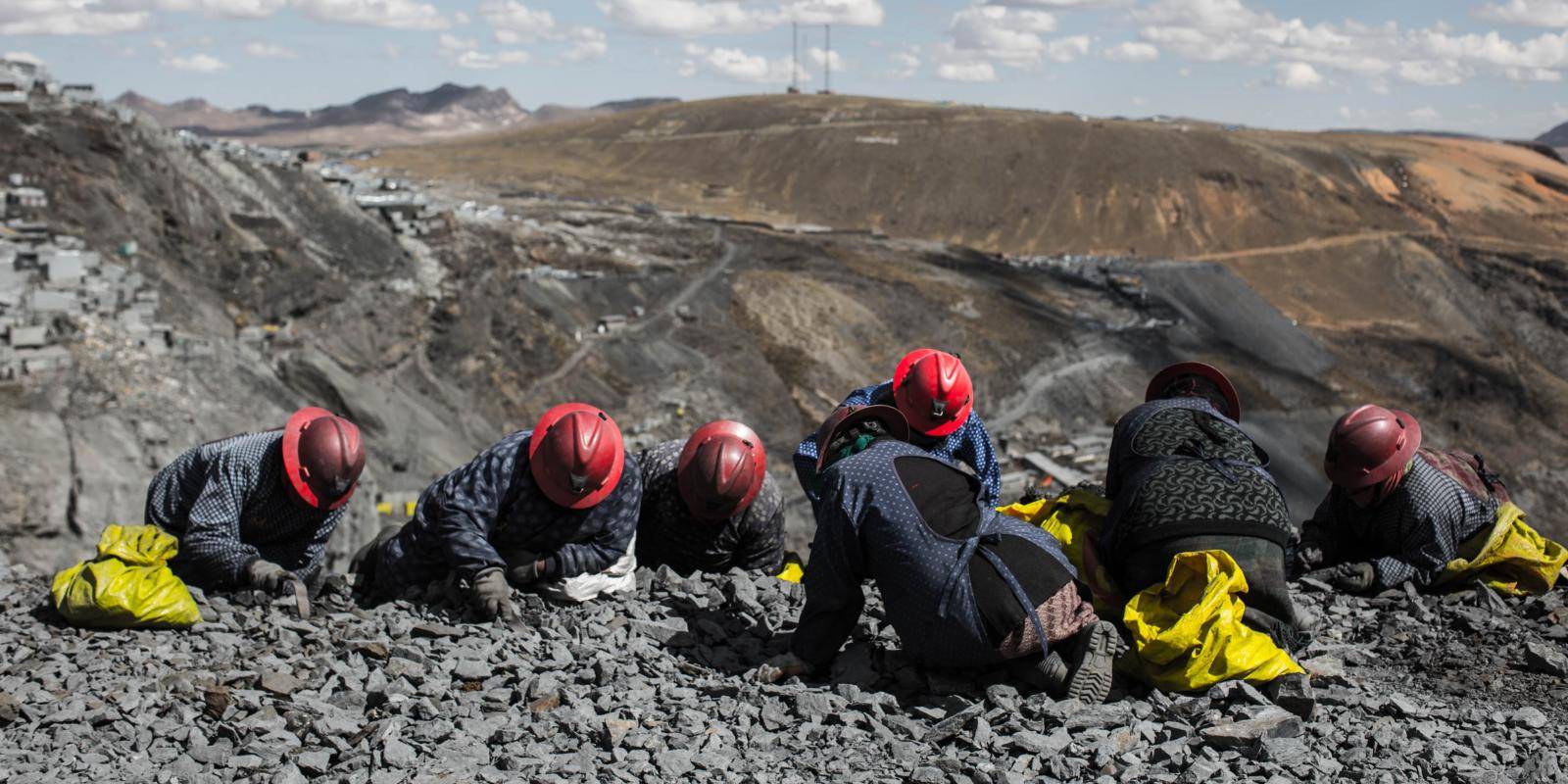 Miners in red helmets search the ground on top of an open pit mine.