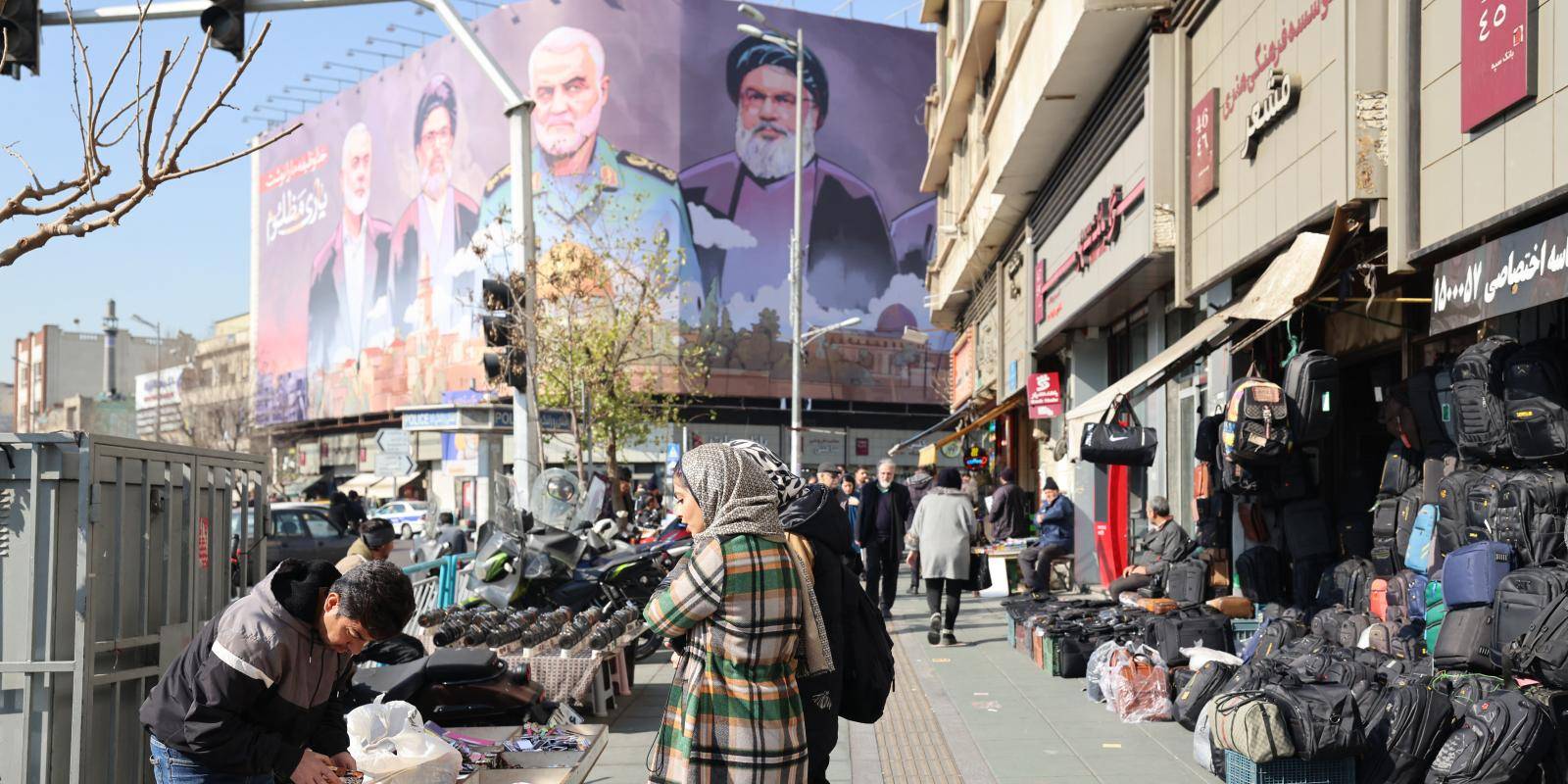 A woman dressed in a winter coat and headscarf looks at a market stall. Above her, a billboard shows the images of four people looking down.