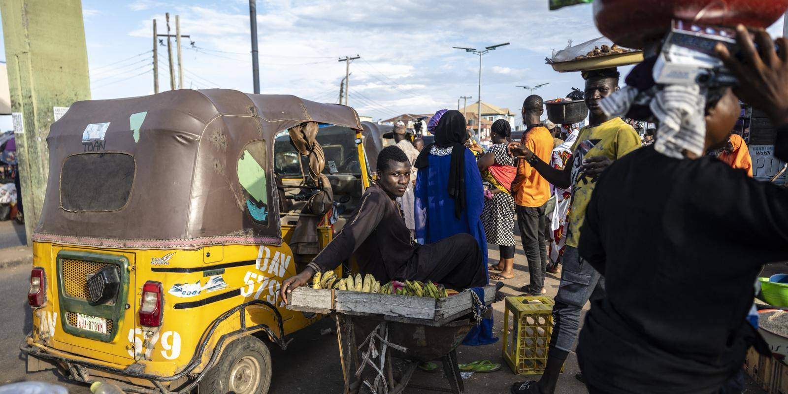 A man sits on a cart, with bananas on a wooden tray in front of him. Behind him, men and women stand between market stalls and yellow-painted vehicles.