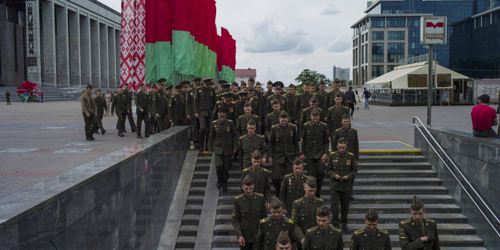 Soldiers in green military dress uniform decend a staircase from an open square. Red and green Belarusian flags can be seen to the left.