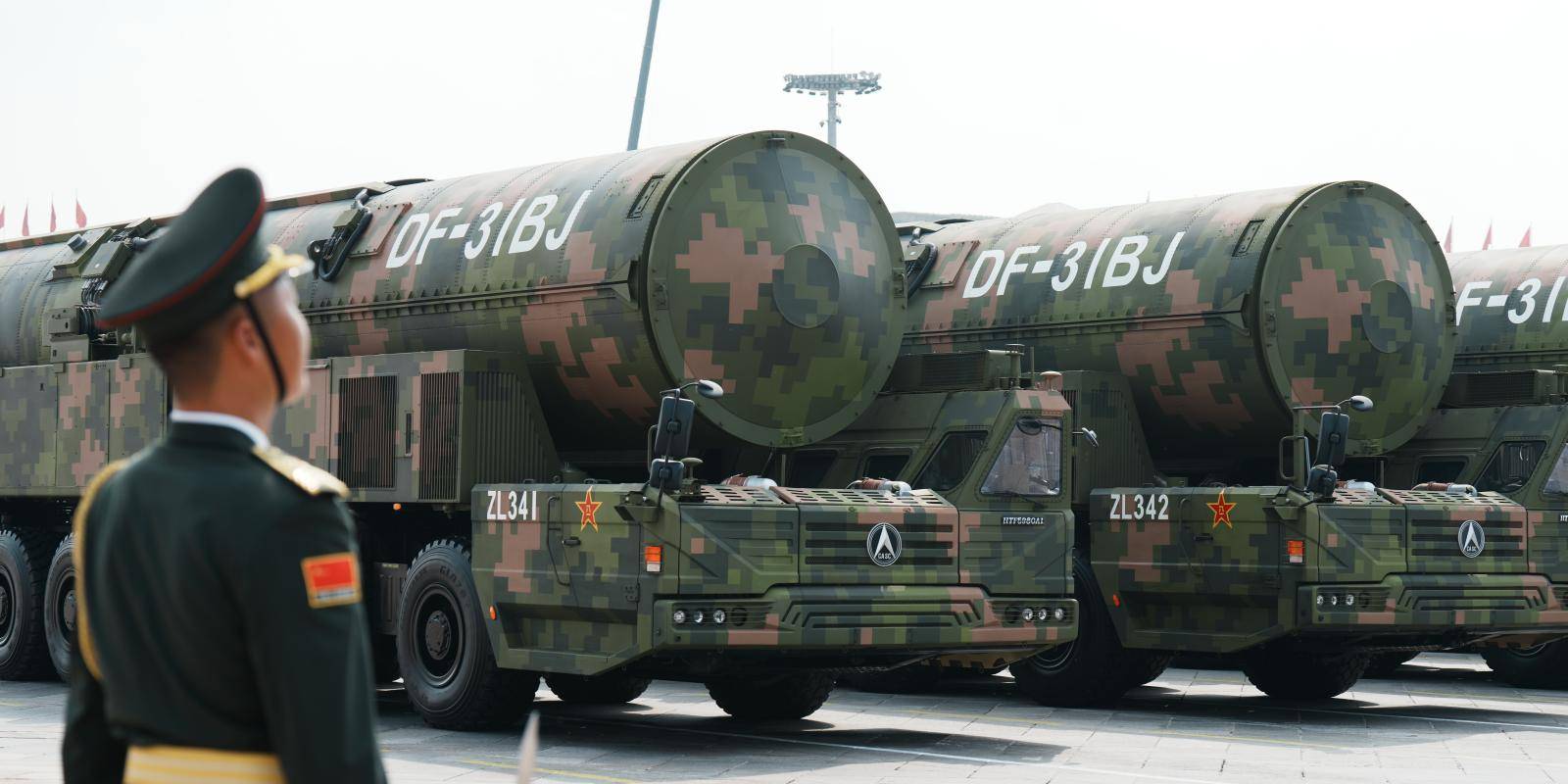 To the left of the frame, a soldier in green dress uniform stands to attention as three trucks carrying missiles pass by in formation..