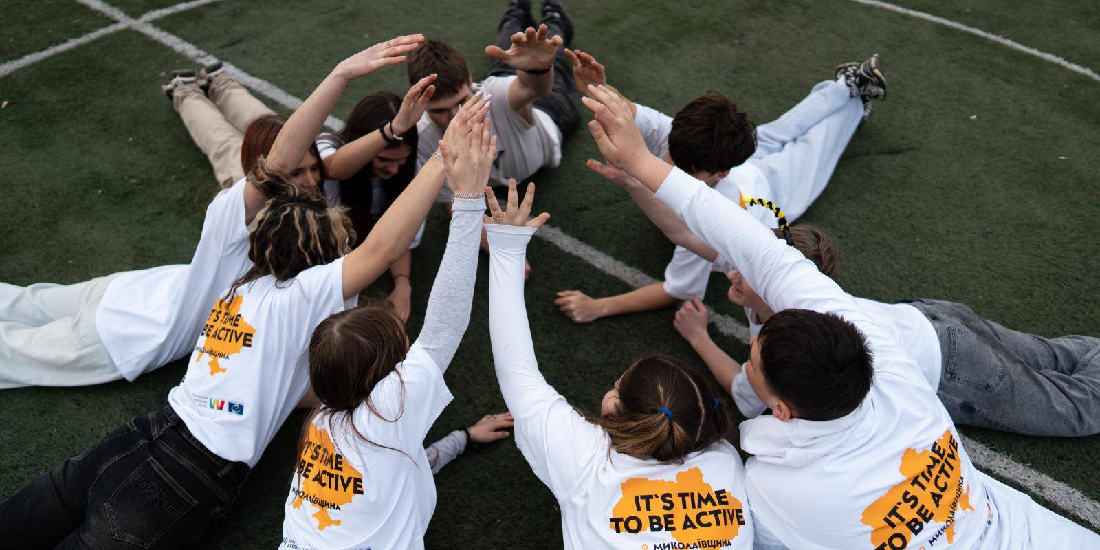 A group of teenagers in white t-shirts lay on the ground, forming a circle and raising their hands together.