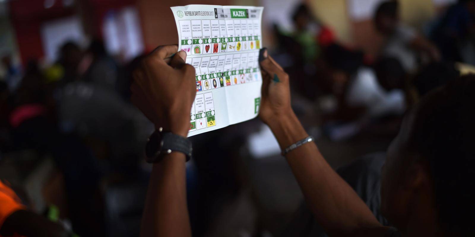 A person holds a white and green sheet of paper with party logos and tick boxes.