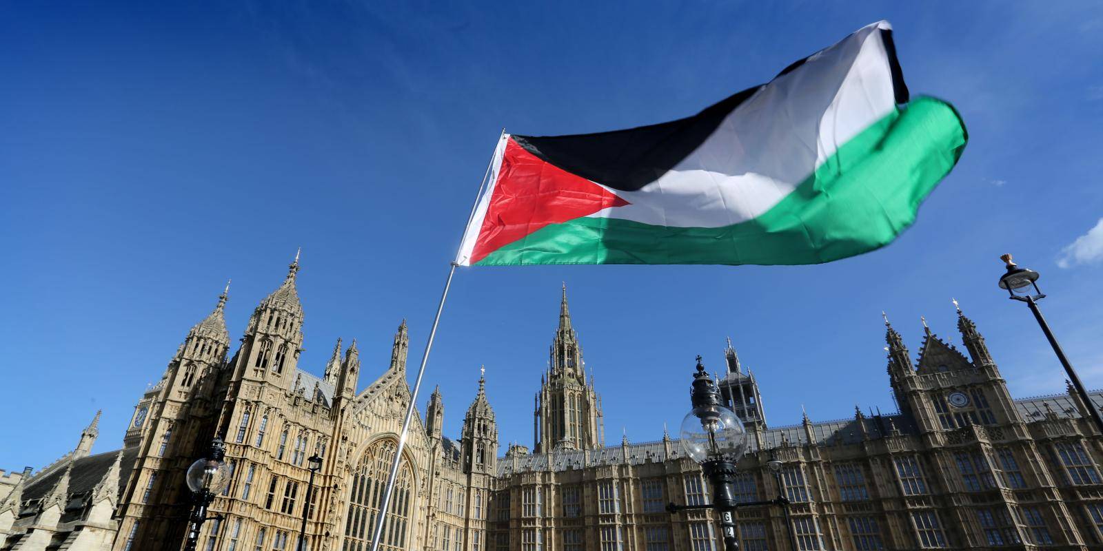 A green, red, black and white Palestinian flag against a blue sky in front of the gothic parliament building in London, UK