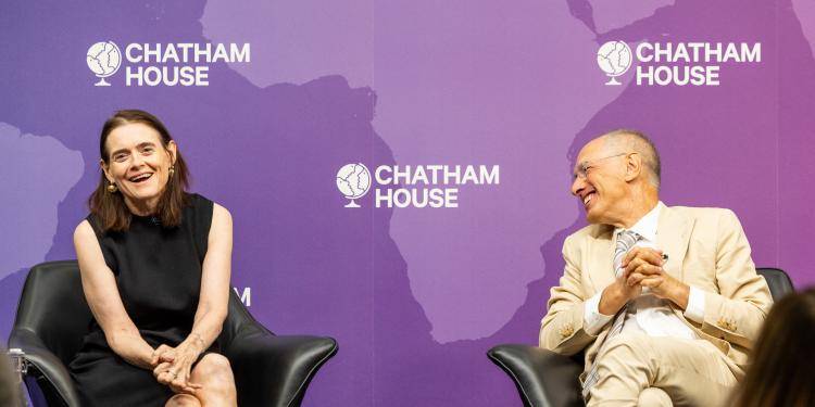 Bronwen Maddox in a black dress and Sir Michael Moritz in a beige suit laugh together while seated on stage in front of a purple Chatham House backdrop.