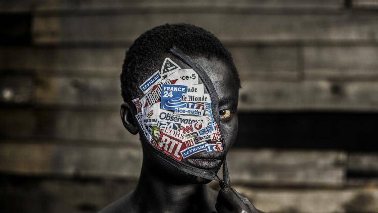 Photo portrait of a woman in the Democratic Republic of the Congo, her left eye towards the camera, her right eye obscured by a Covid-style mask pulled obliquely over her face