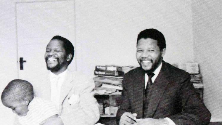 A smiling Oliver Tambo, with toddler on lap, and Nelson Mandela of the ANC at a desk, possibly in 1961