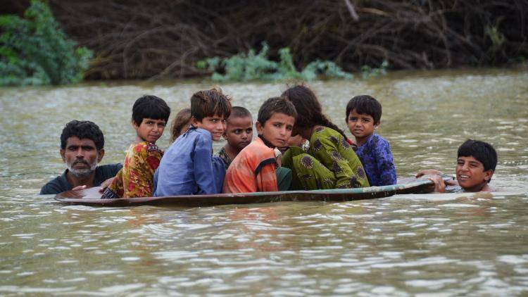 Two men guide seven children in a large plastic container across a flooded area in Pakistan, 2022