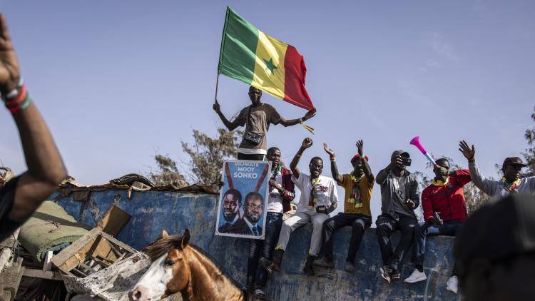 A man stands on a wall waving a Senagalese flag, with other men and a horse in the foreground