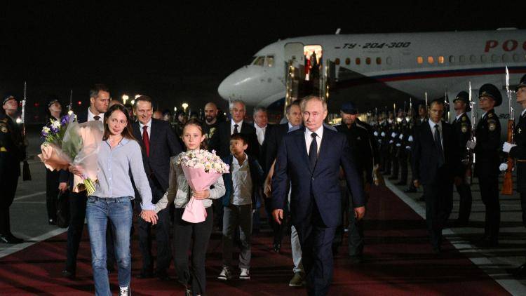 a man in a suit walks alongside  a mother and daughter holding flowers with an airplane behind them