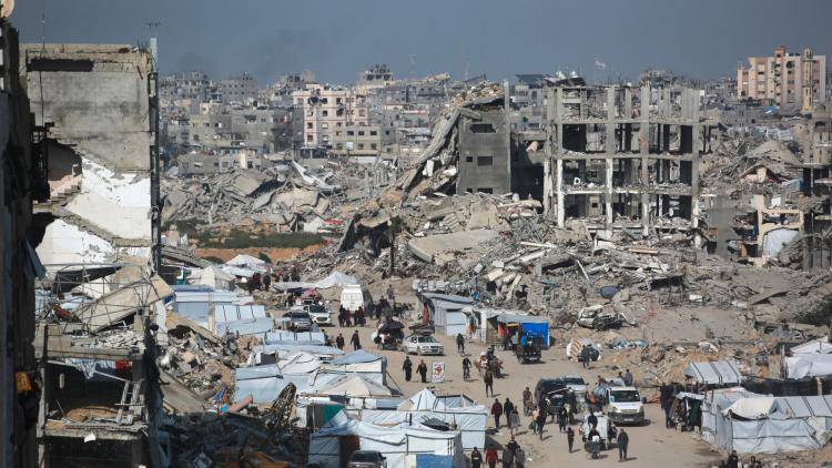 Palestinian vehicles drive through a heavily damaged street in Gaza, lined by tents, overlooked by shelled-out buildings.