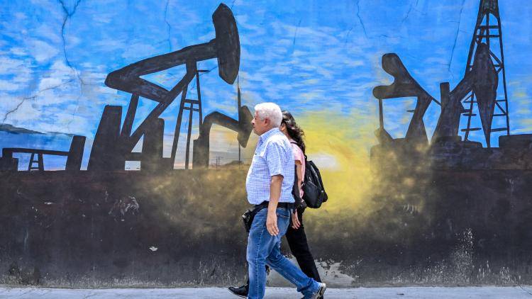 A man and woman walk in front of a blue, black and yellow mural depicting an oil pumps in Caracas, Venezuela
