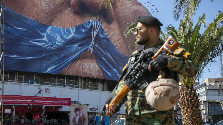 An Iranian police officer stands before a poster in Tehran, depicting President Trump's mouth gagged by a Strait of Hormuz shaped ribbon - May 2026.