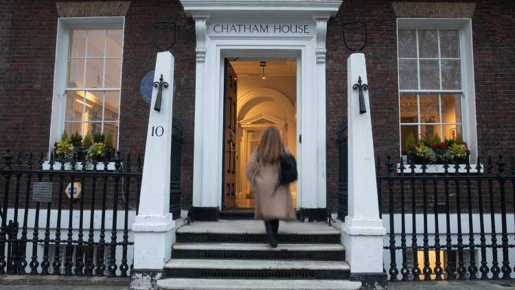     A front exterior view of the brick Chatham House building entrance. A woman with long hair wearing a tan coat walks up the short stone steps toward the open front door, which reveals a brightly lit white interior hallway. The white doorframe is topped with the words 'CHATHAM HOUSE,' flanked by white stone pillars marked with the number '10' and black iron railings.