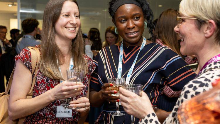 Three women holding crystal wine glasses stand closely together, smiling and chatting at an indoor evening event. The woman on the left wears a red patterned dress and a "DELEGATE" badge. The woman in the center wears a striped navy dress, a black headband, and a blue and white Chatham House lanyard. The woman on the right, seen in profile, wears glasses and an animal-print top. Other attendees are softly blurred in the background.