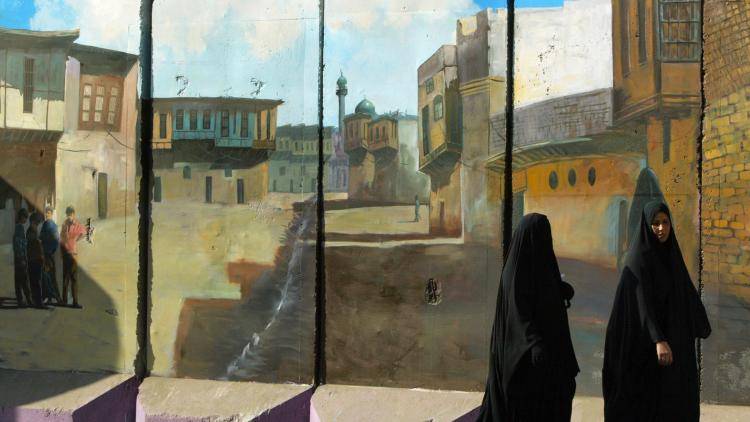 Iraqi women walk past a mural decorating a concrete blast wall in Baghdad