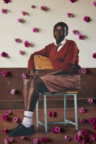 Portrait of a schoolgirl in uniform sitting sideways on a chair, the wall behind her decorated with mauve flower petals