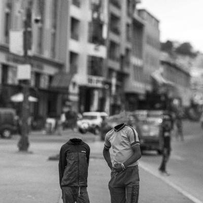 Black-and-white urban street scene in Madagascar with two young boys posing in the foreground, their heads digitally removed from the image