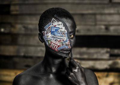 Photo portrait of a woman in the Democratic Republic of the Congo, her left eye towards the camera, her right eye obscured by a Covid-style mask pulled obliquely over her face