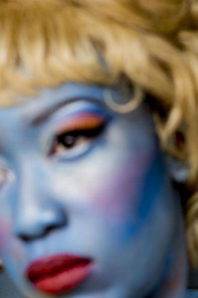Blurred close-up portrait of a woman with a face garishly made up in heavy blue foundation, red lipstick, orange eyelids and blonde hair