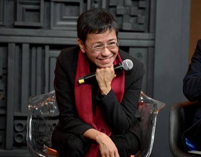 Maria Ressa sits in a chair, smiling and holding a microphone