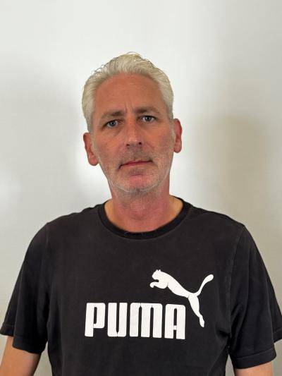 man in black t-shirt stands in front of a white wall looking into the camera