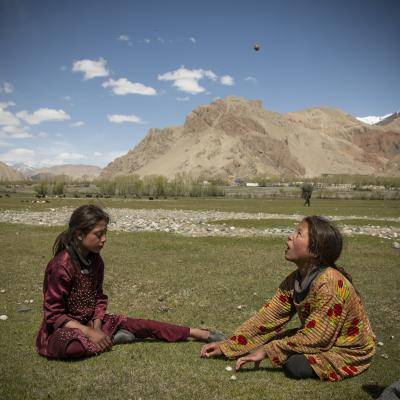 two young girls sit on the grass playing a game with stones with mountains behind them