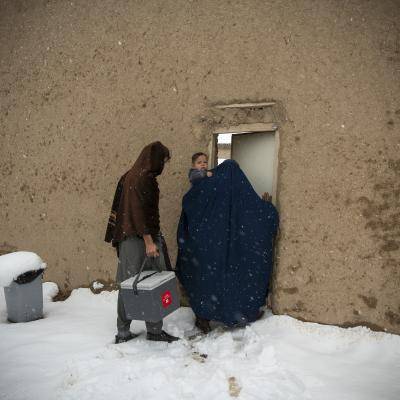 a woman covered in a head scarf holding a baby opens a door to a building, with a man standing next to her in the snow. 