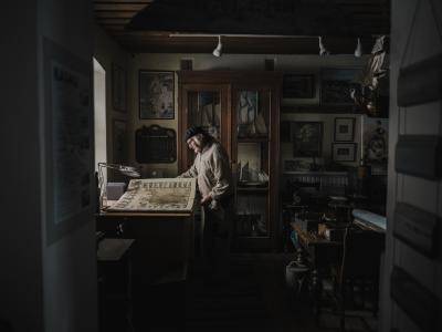 older man stands in dark room in a maritime museum looking at a map on a desk by the window