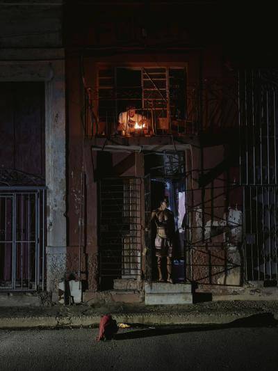 A man cooks by a fire on his balcony while the street below is in darkness